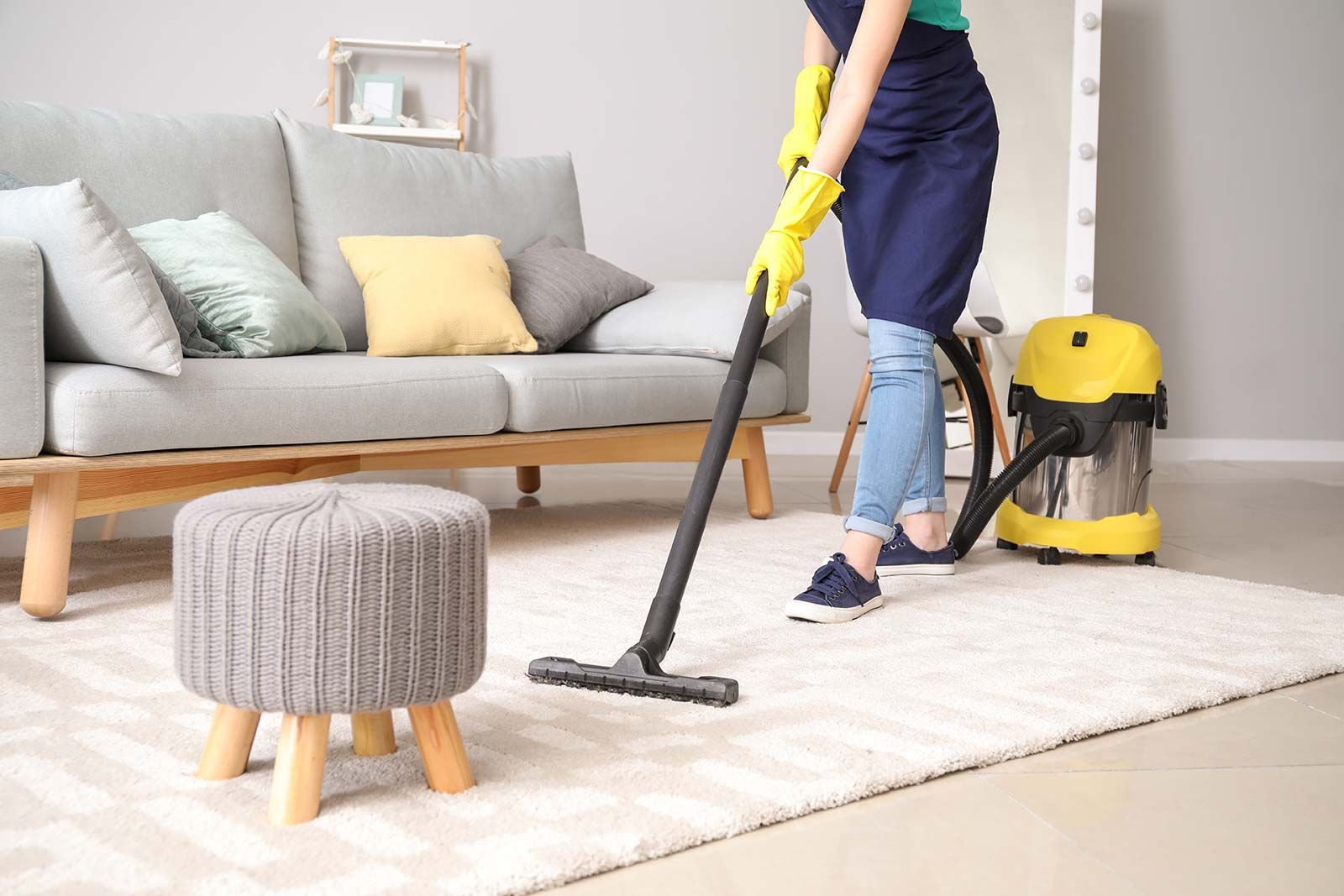 Person vacuums a white rug in a living room, wearing gloves and an apron, near a gray couch and a small ottoman.