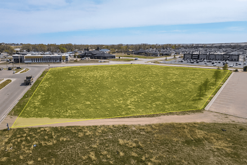 Aerial view of a vacant, grassy lot with commercial buildings in the background under a blue sky.