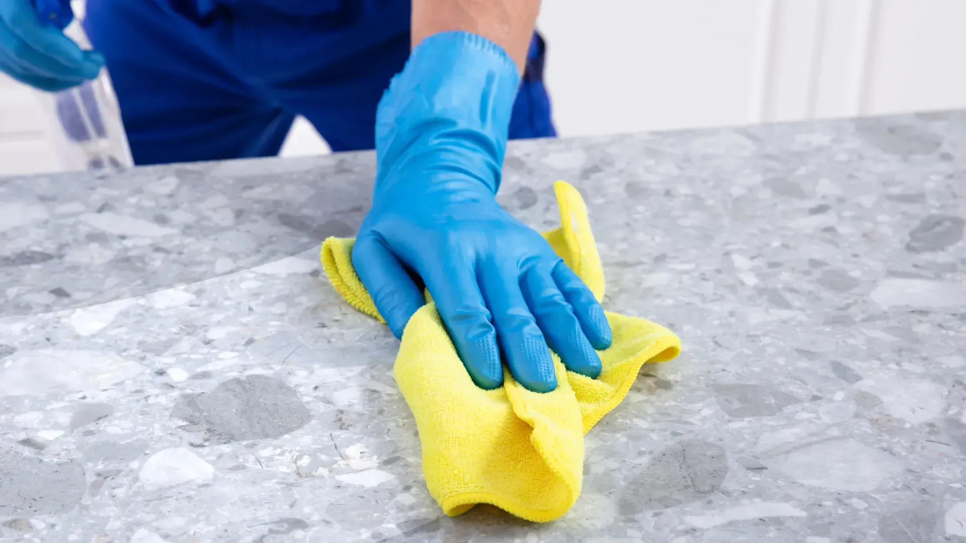 Blue-gloved hands wiping a gray countertop with a yellow cloth