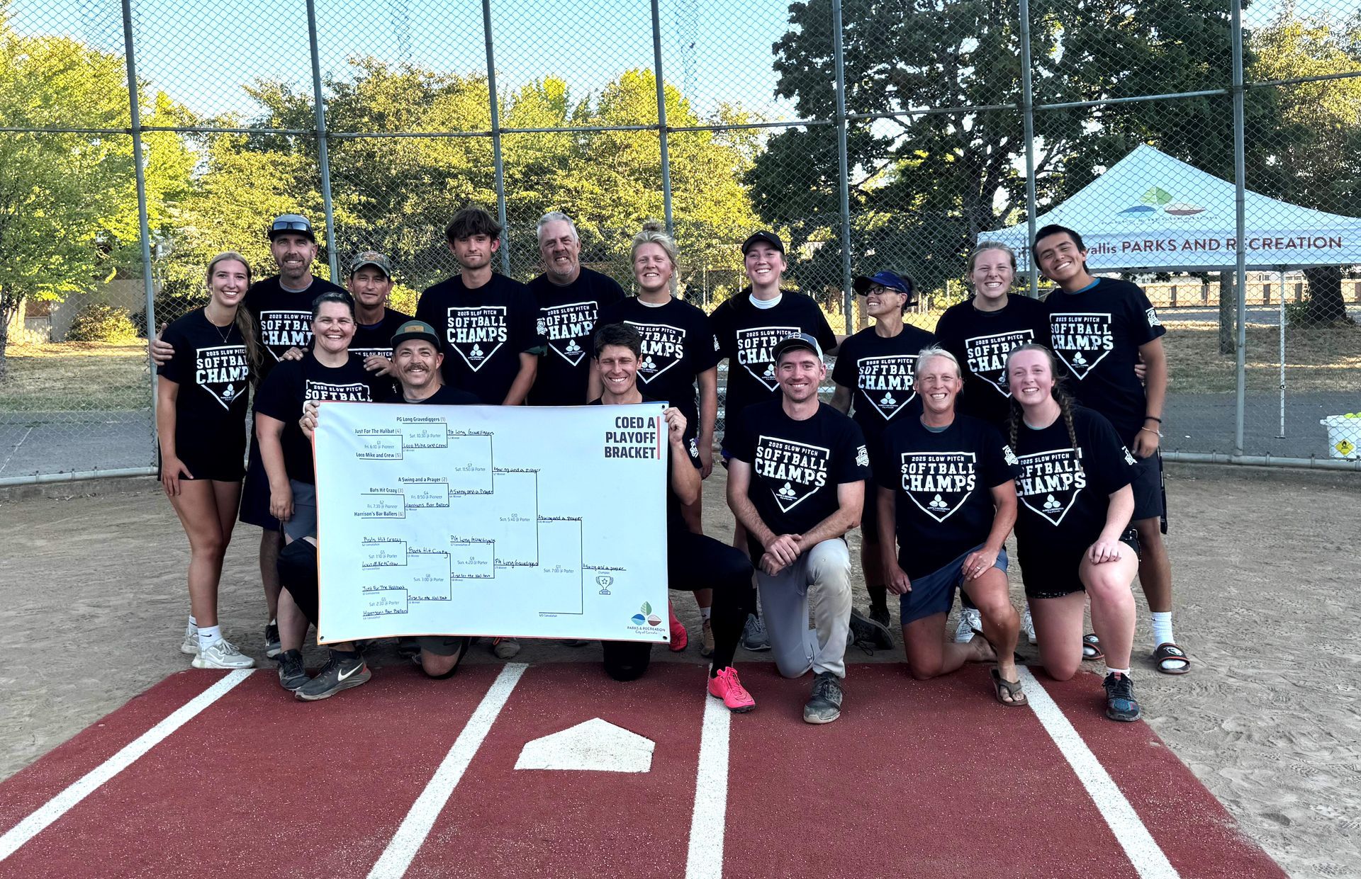 A softball team wearing matching black shirts poses for a photo on a dirt field, holding a tournament bracket sign.