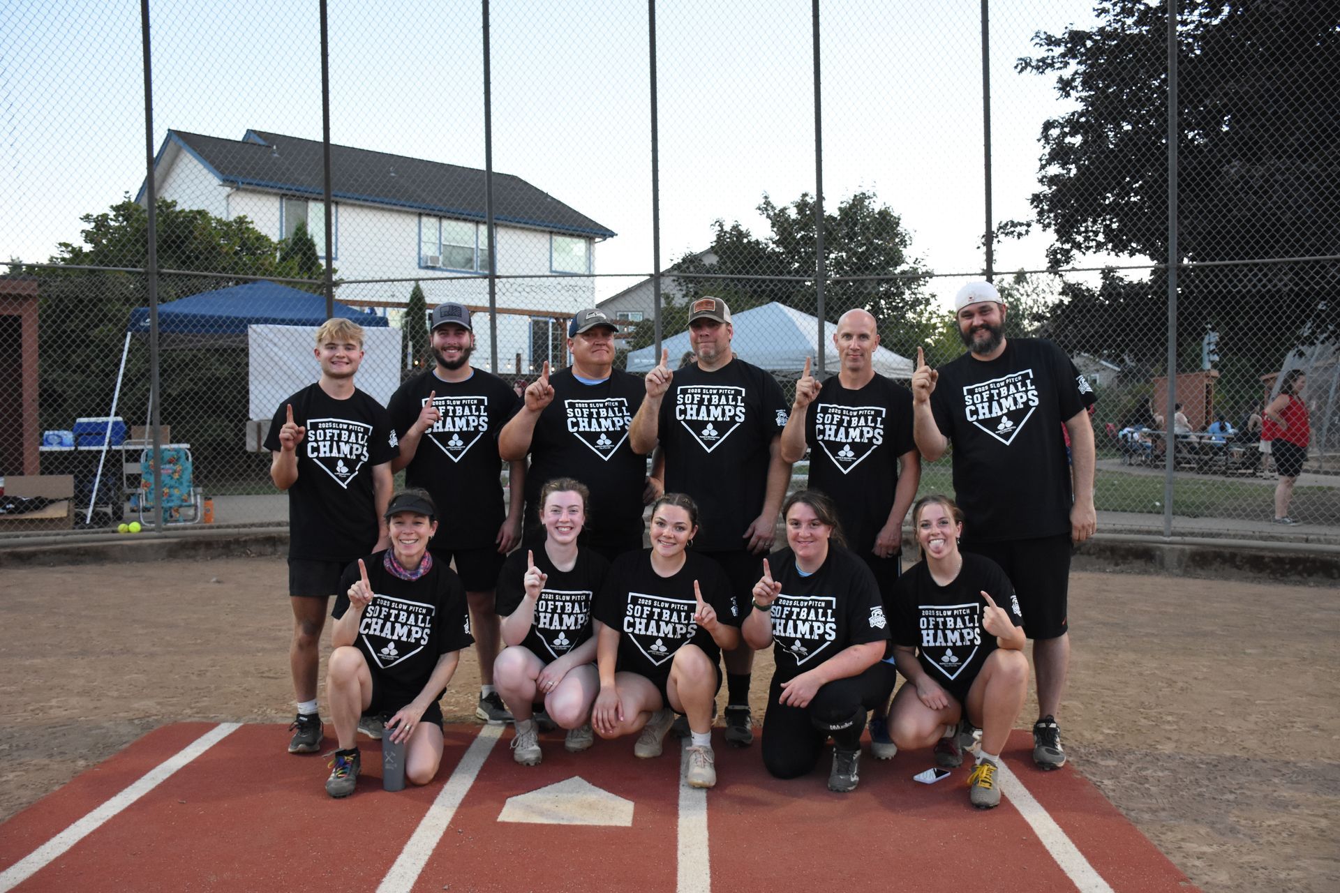 A softball team wearing matching black shirts poses for a photo on a dirt field, holding up one finger in celebration.