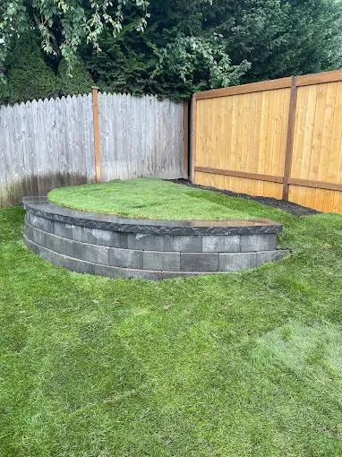 A curved retaining wall made of gray blocks encloses a patch of grass. A wooden fence is behind.
