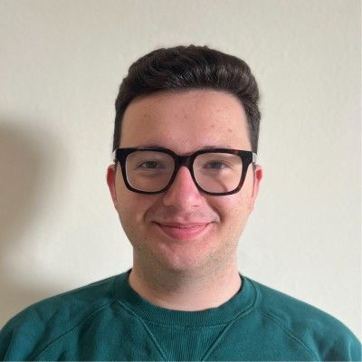 Un uomo con gli occhiali e un maglione verde sorride alla telecamera