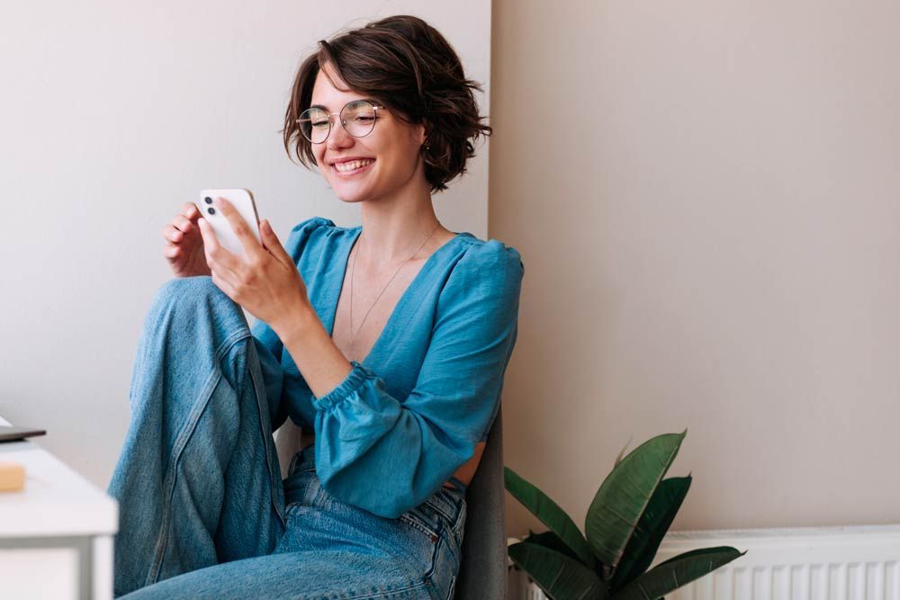 Woman in blue top and jeans smiles while using a phone, sitting near a plant.