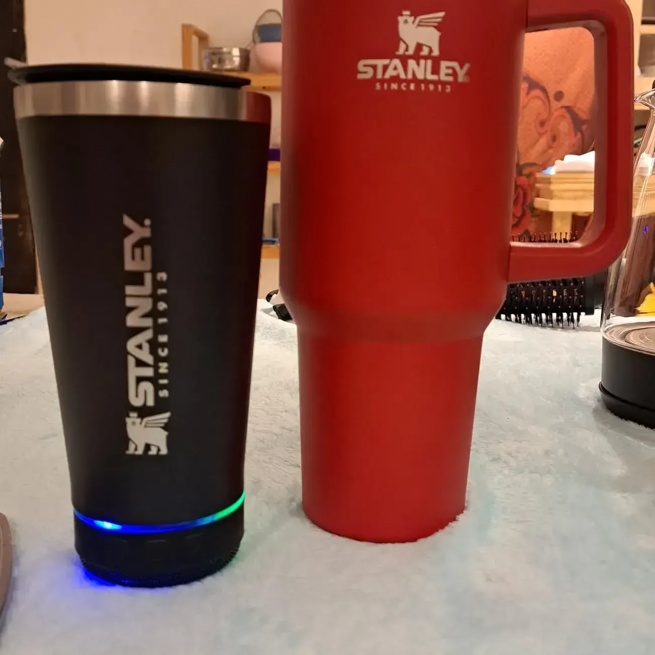 Dos vasos Stanley: uno negro con luz azul/verde y otro rojo con asa.