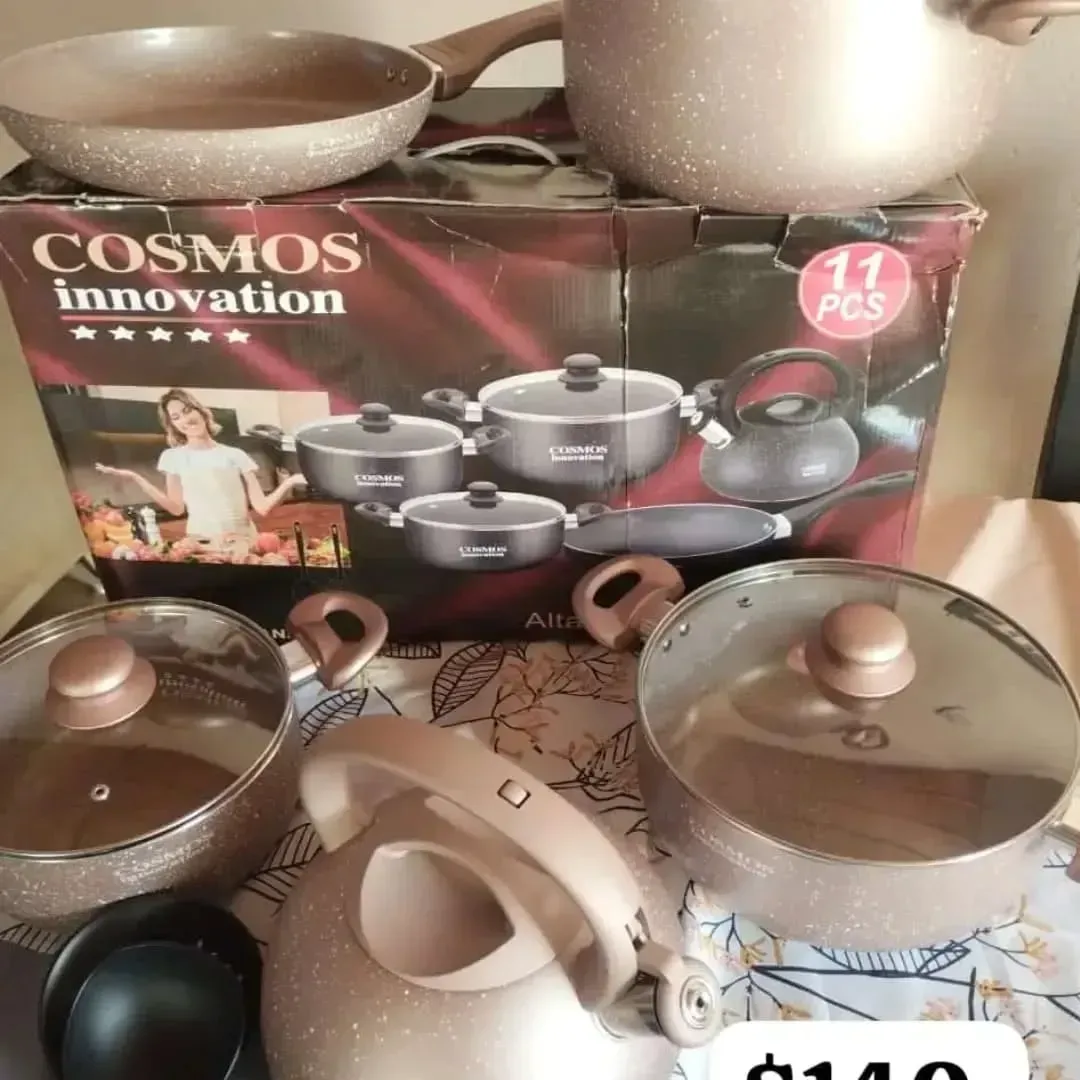 Un juego de utensilios de cocina de 11 piezas en caja. Sartenes, ollas y una tetera se presentan en colores dorado y crema.