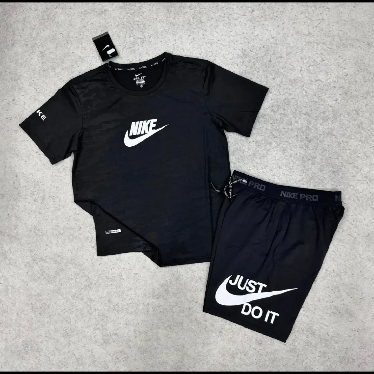 Conjunto de camiseta y pantalones cortos Nike negros con logotipos blancos sobre superficie gris.