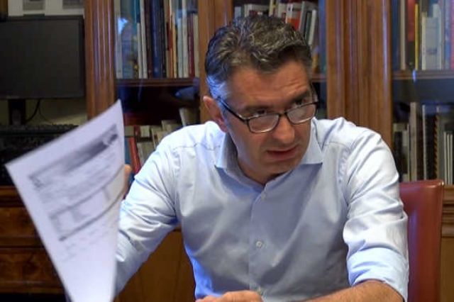 Un uomo con gli occhiali è seduto alla scrivania con in mano un pezzo di carta.