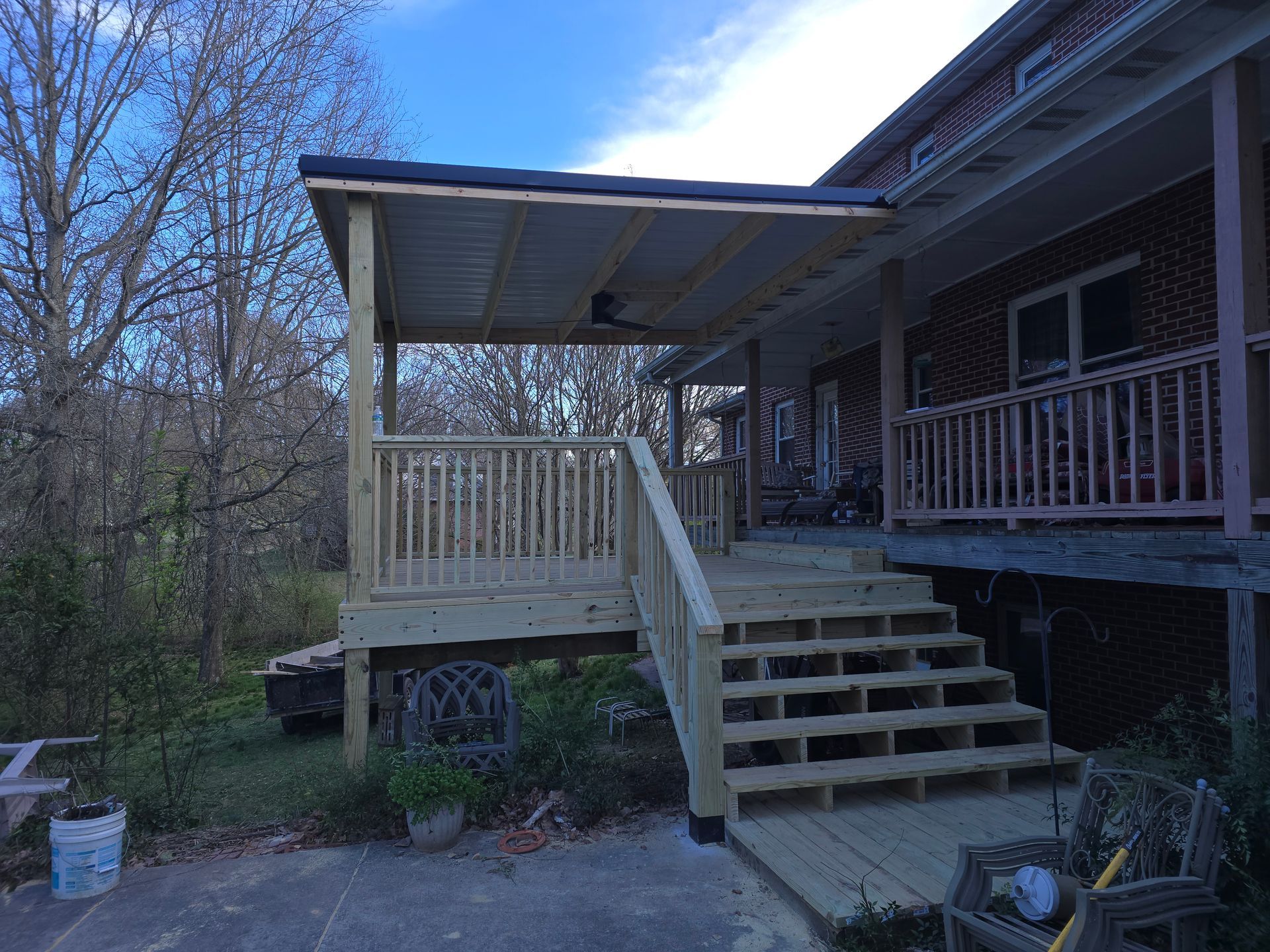 Wooden porch with steps, railing, and covered roof extending from a brick house. Trees in the background.