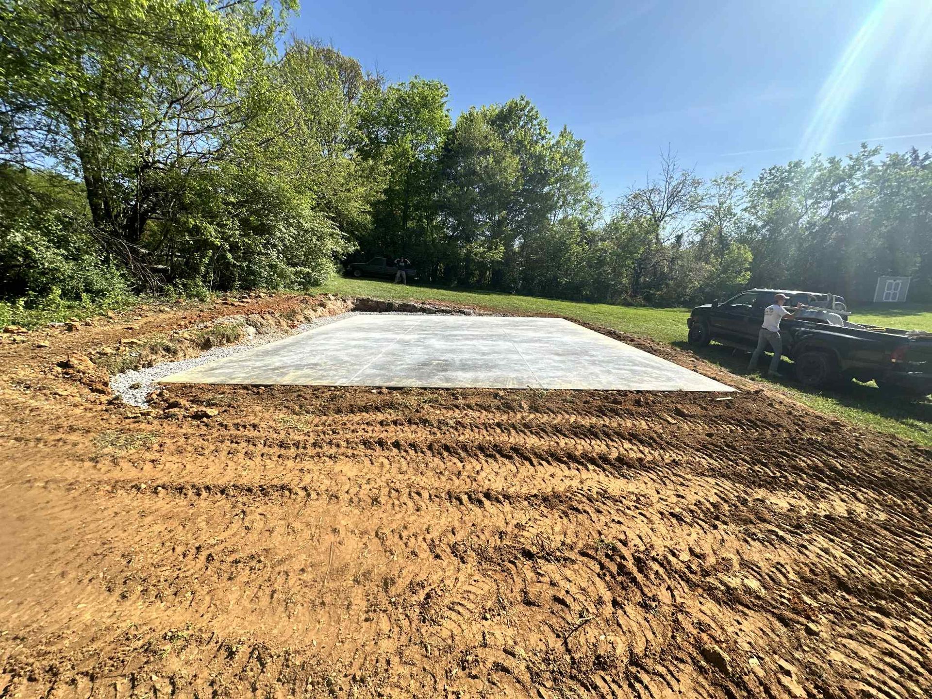 Concrete slab in a cleared area with a tractor in the background; dirt and trees surround.