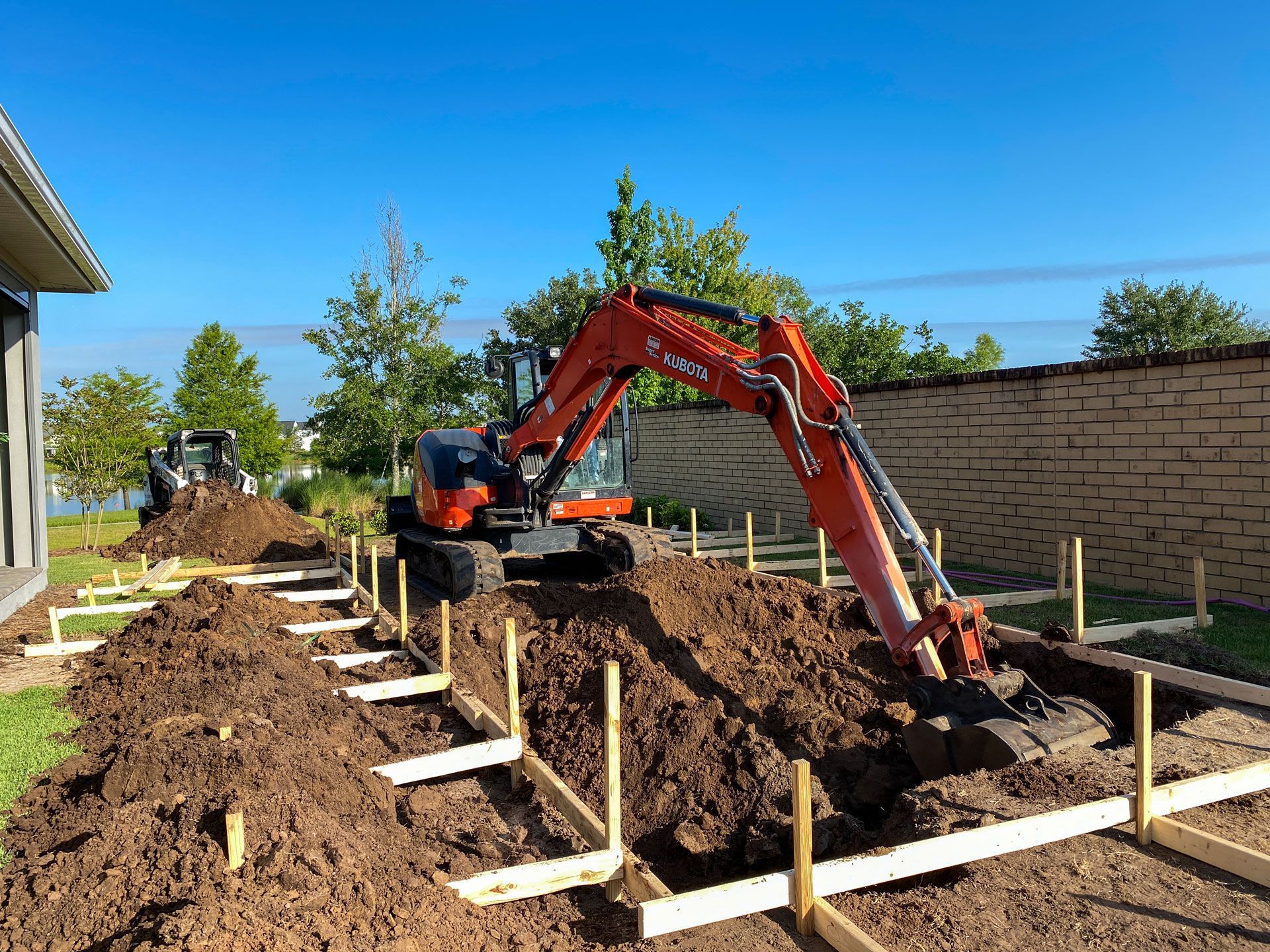 An excavator digs in a dirt plot framed with wooden stakes, near a house and wall under a blue sky.