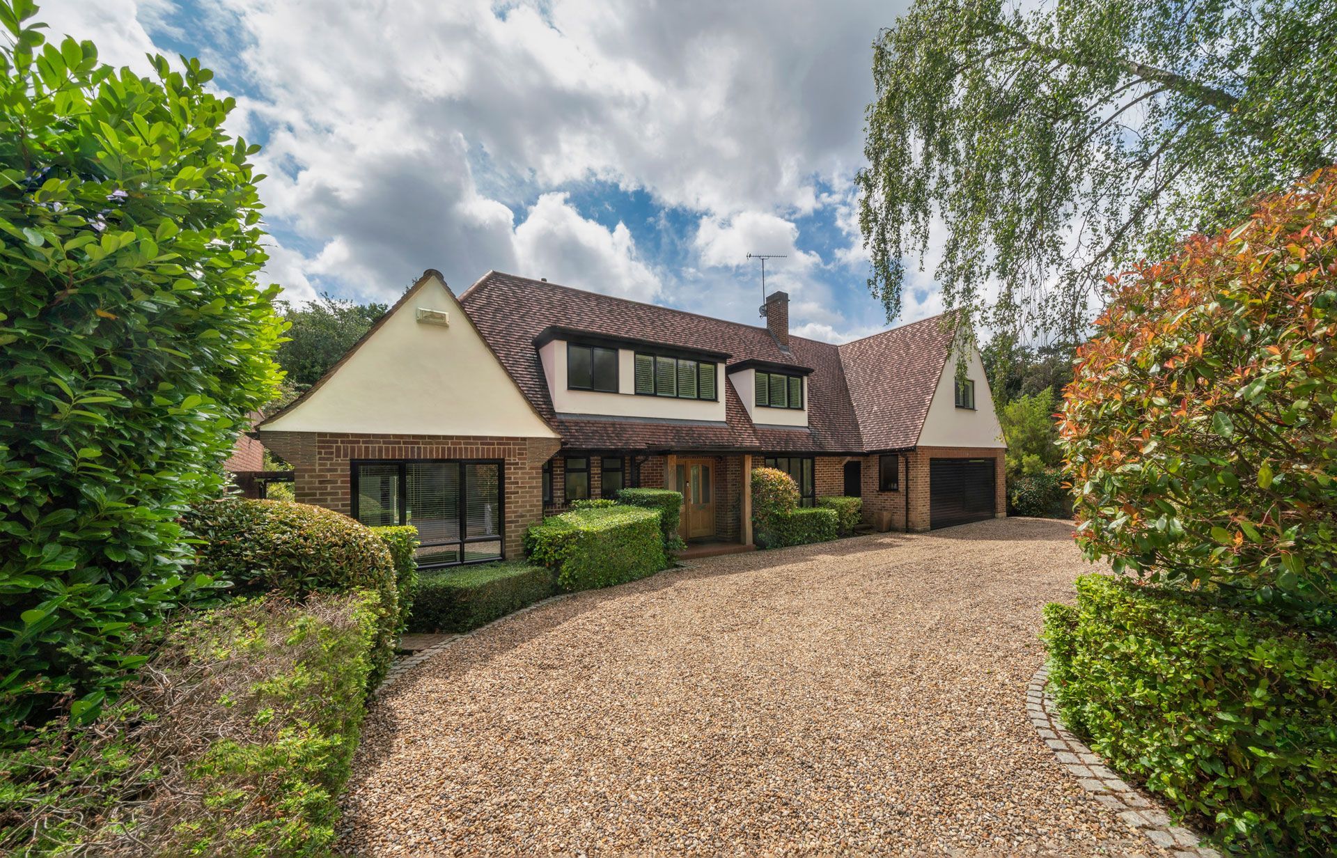 House with brown roof and driveway surrounded by greenery under a cloudy sky.