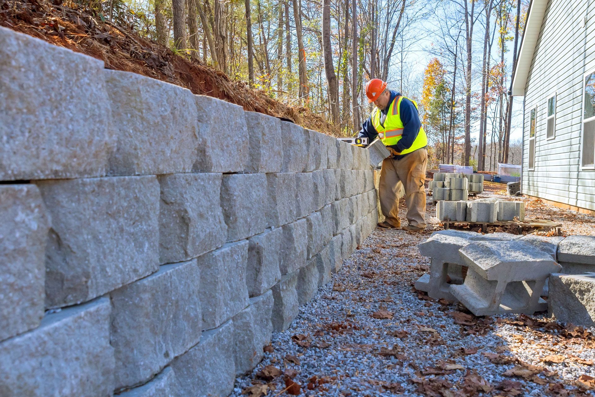 A construction worker builds a retaining wall from concrete blocks next to a house.