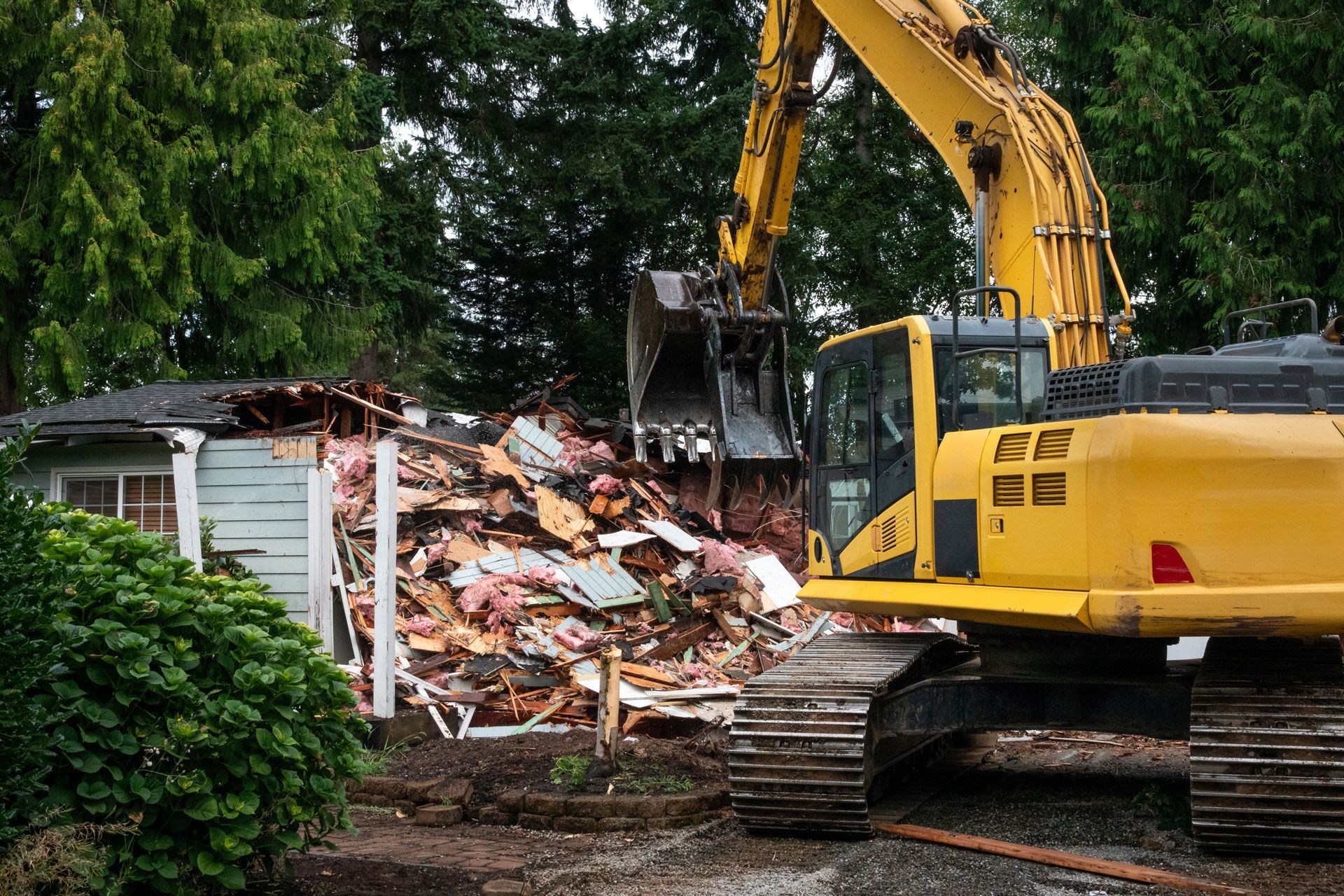 Yellow excavator demolishing a small blue building, surrounded by trees.