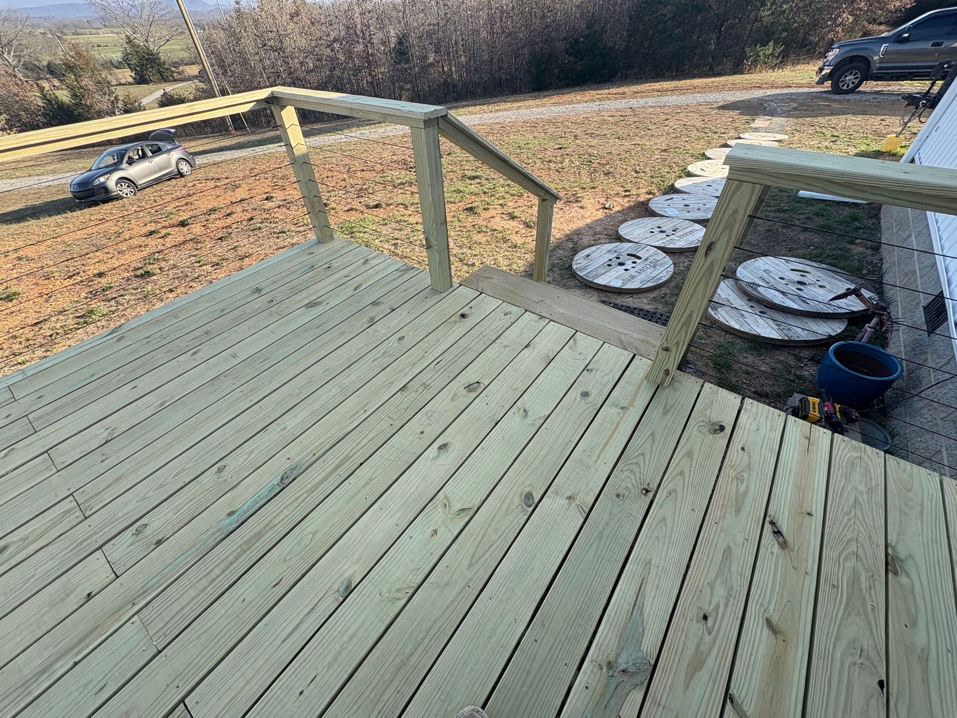 Wooden deck with handrails and steps, overlooking a yard and road with a car.