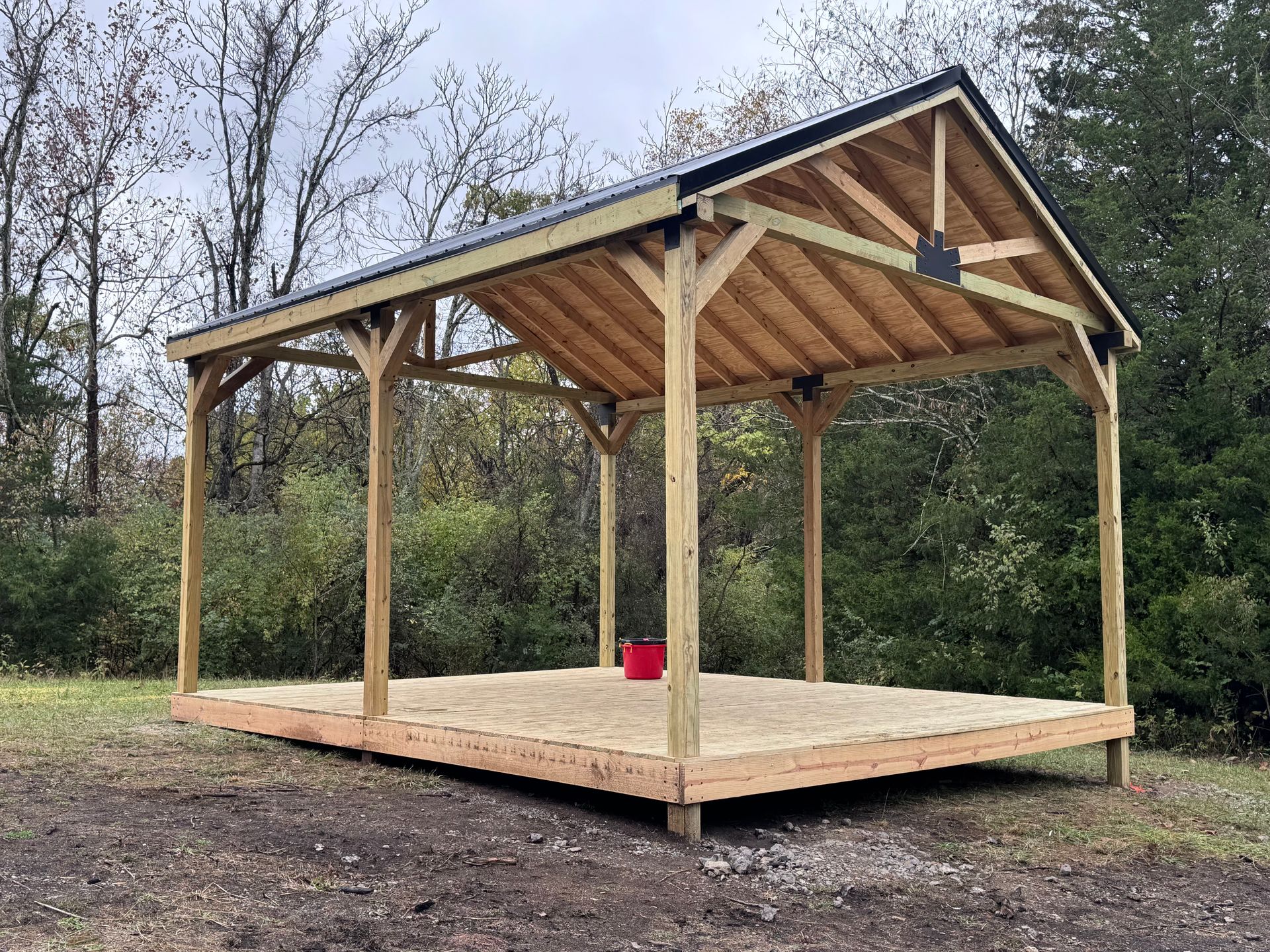 Wooden open-air shelter on a wood platform with a black roof and open sides, set in an outdoor wooded area.