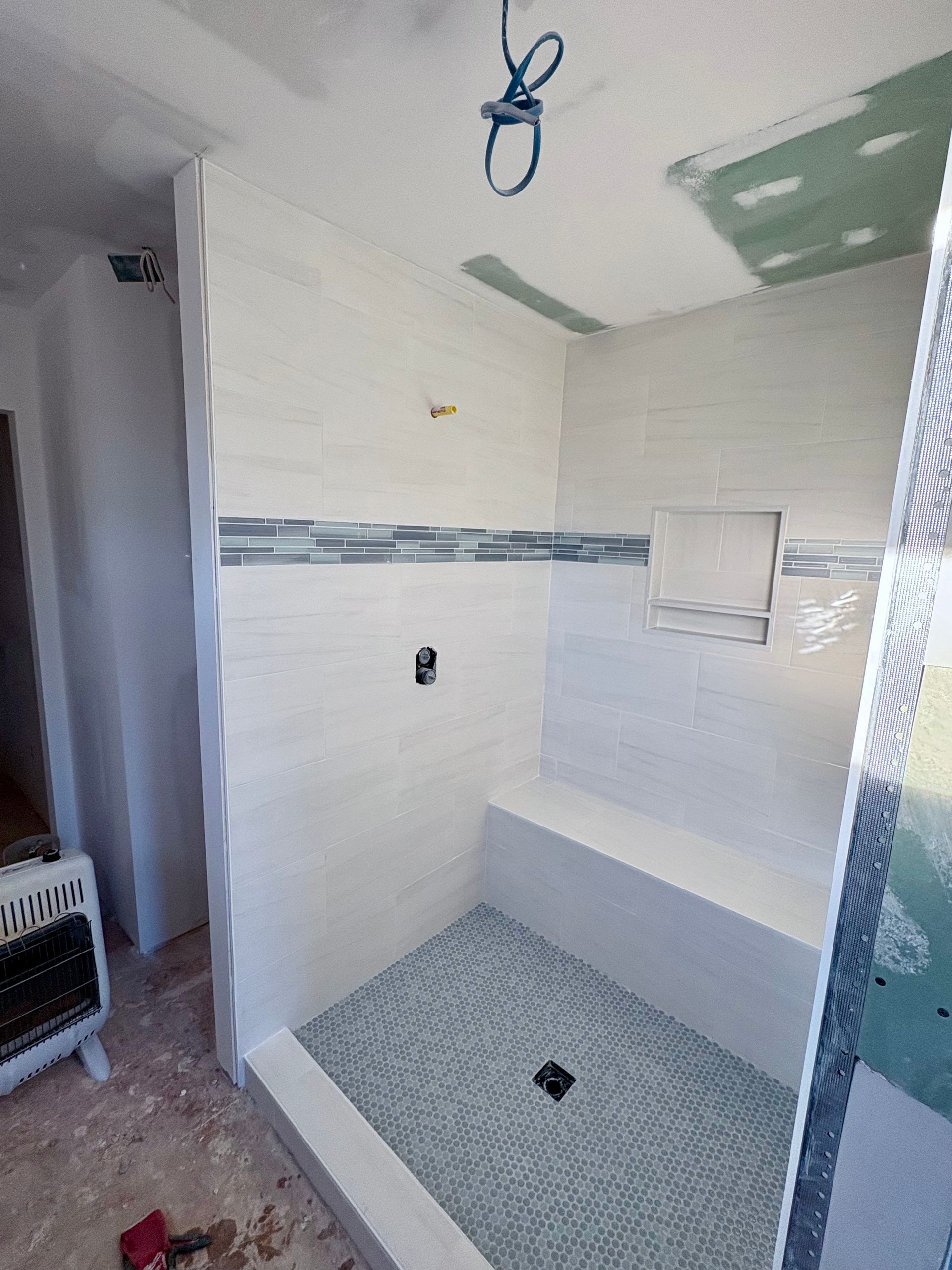 Partially tiled shower in progress; white tiles, blue accent, built-in seat, gray floor, incomplete ceiling.