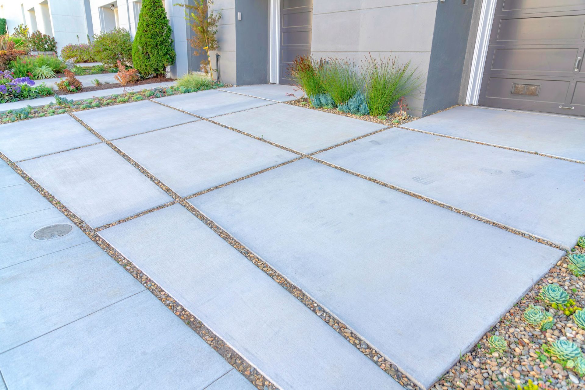 Concrete driveway with gravel borders and greenery alongside a modern home with gray garage doors.