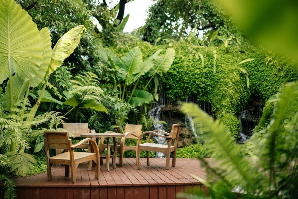 A wooden deck with a table and chairs in a lush green garden with a waterfall in the background.