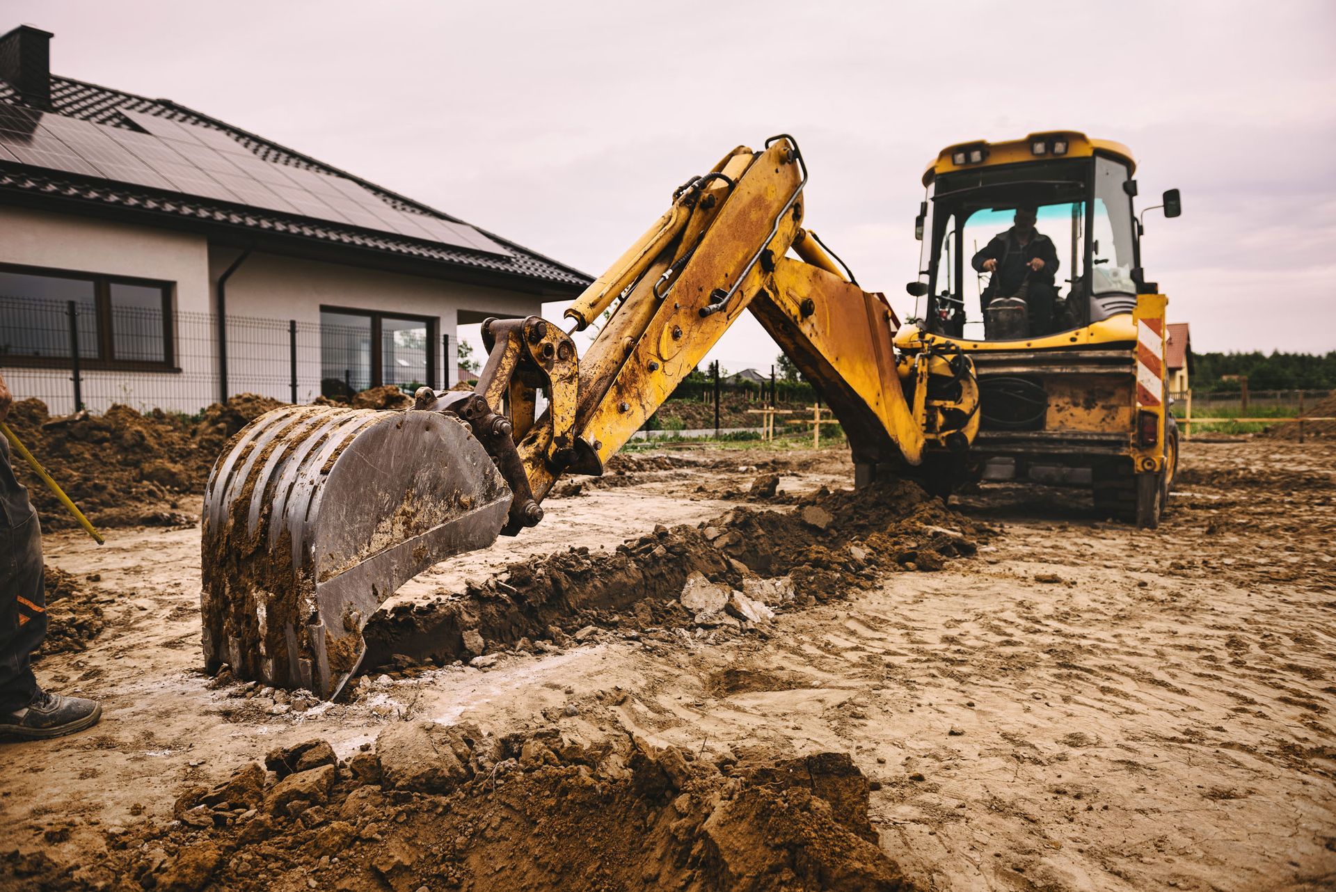 Yellow excavator digging a trench near a house under construction; a worker operates the machine.