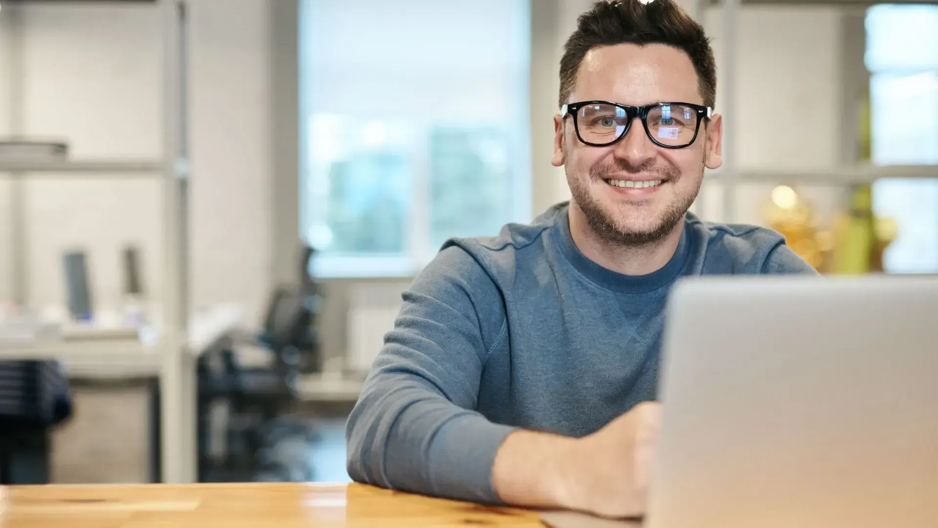 A man wearing glasses is sitting at a desk in front of a laptop computer.