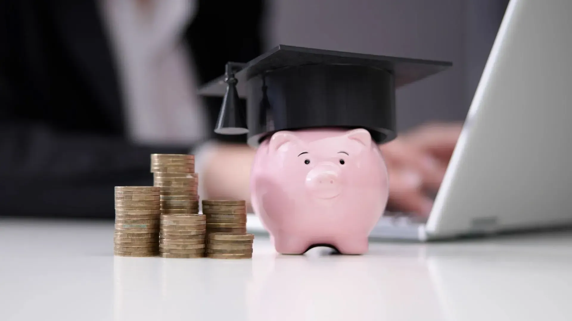 A piggy bank wearing a graduation cap is sitting next to stacks of coins.