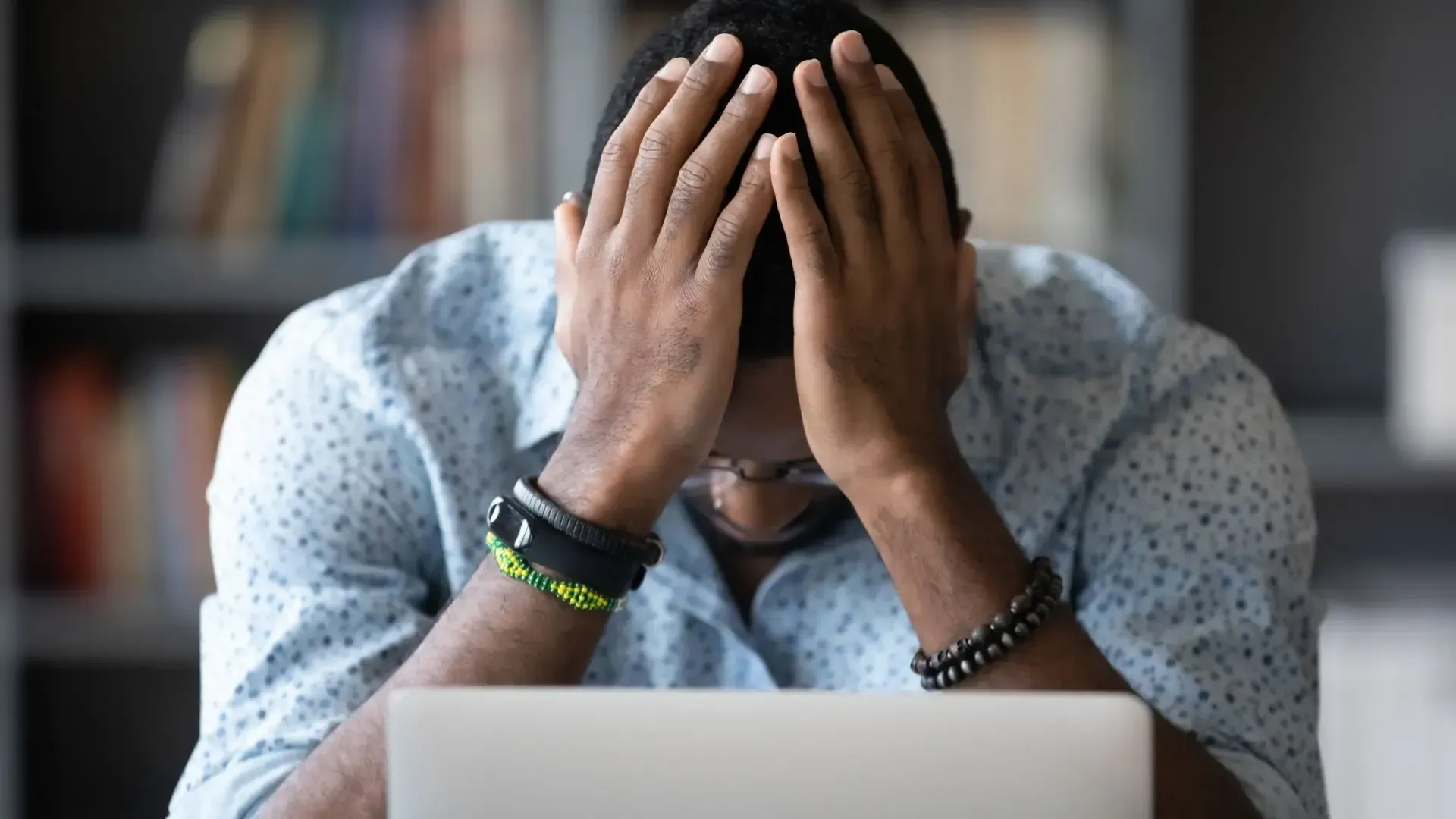 A man is sitting in front of a laptop computer covering his face with his hands.