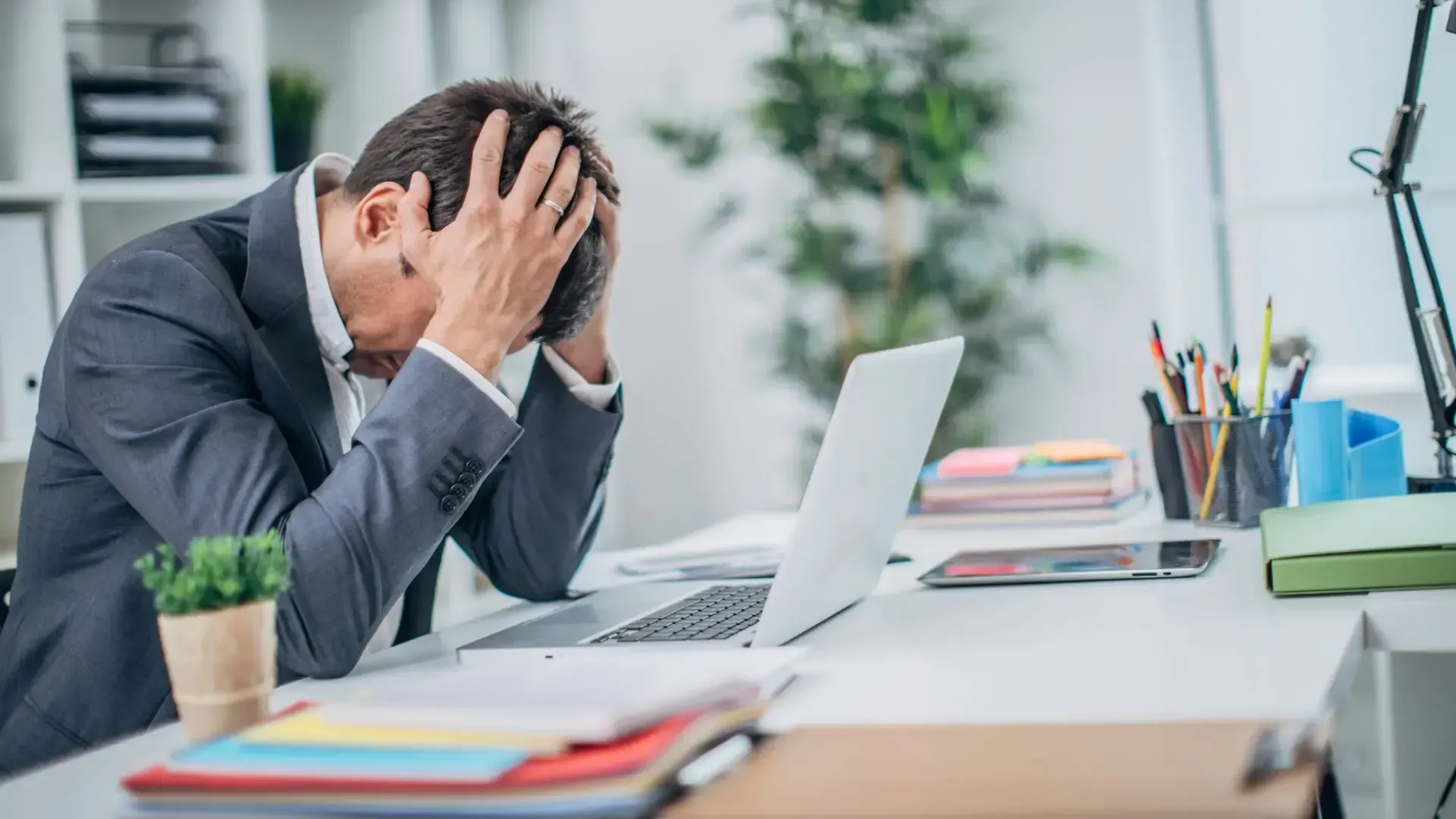 A man in a suit is sitting at a desk with his head in his hands in front of a laptop computer.