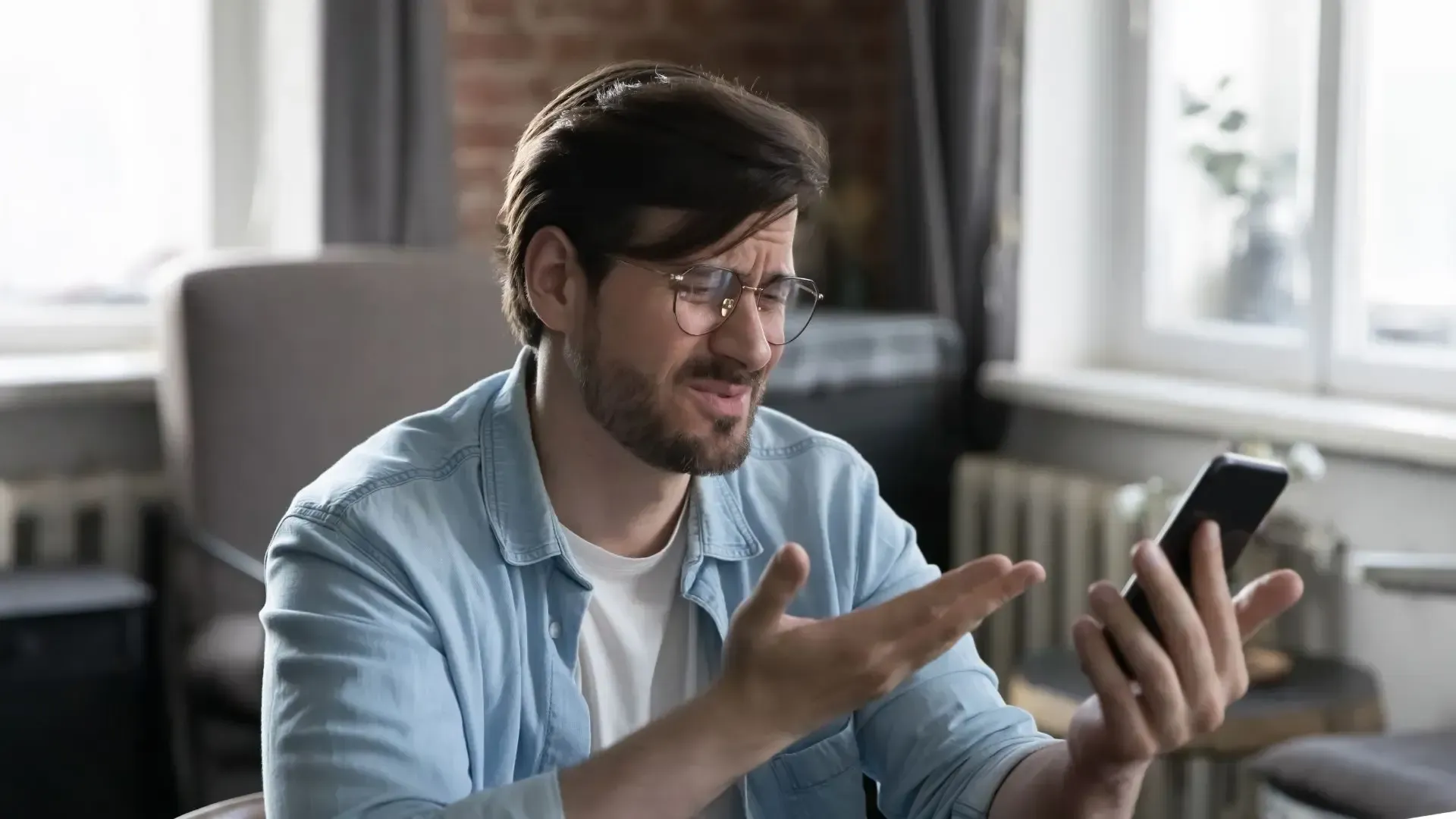 Man with glasses looking frustrated at his phone, gesturing with his hand in a room with a brick wall.
