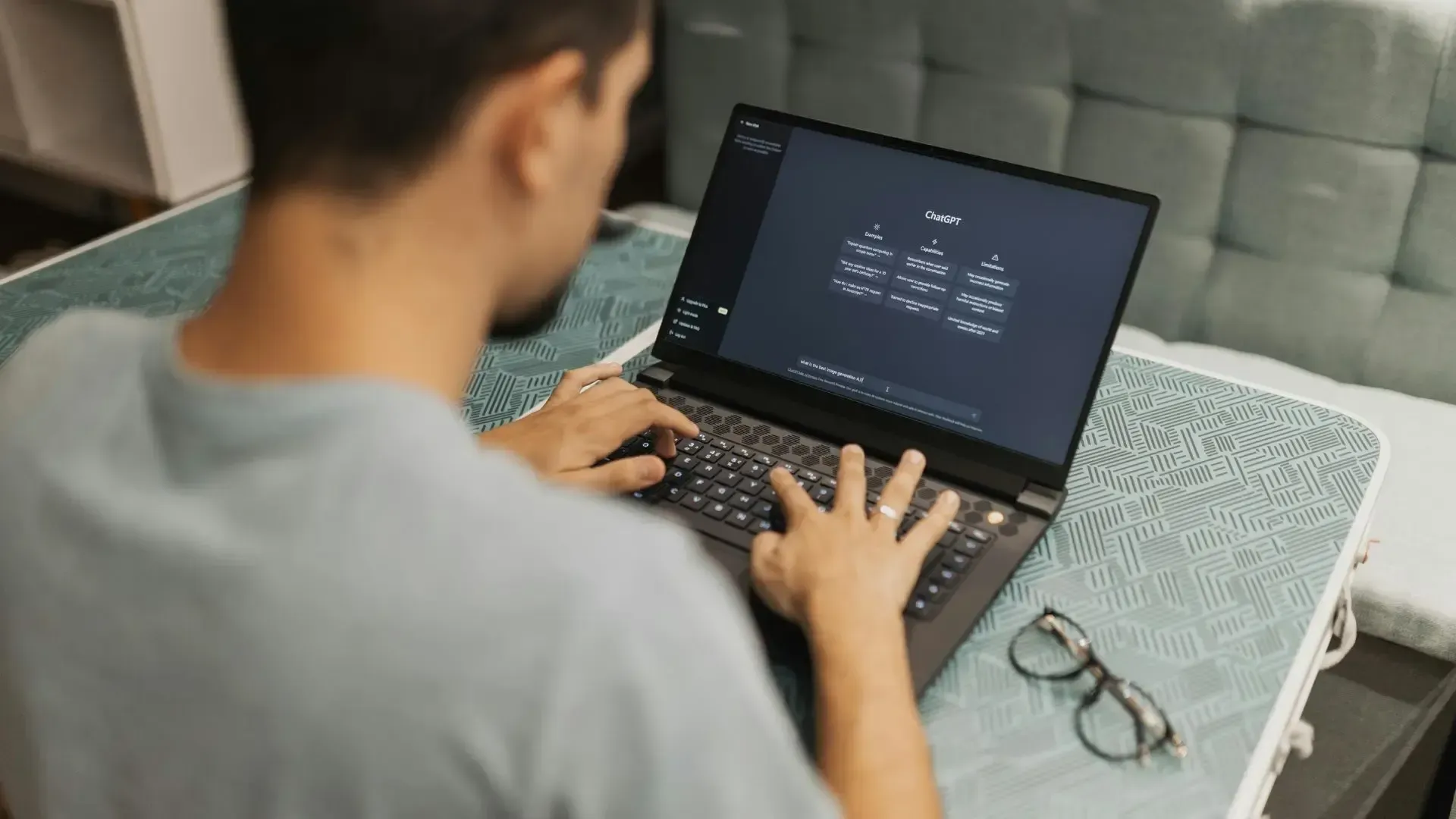 Person seated at a desk, using a laptop. Screen shows a dark user interface, glasses nearby.