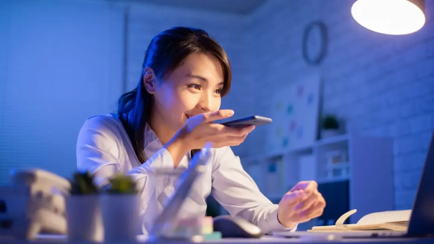 Woman in a lit office, holding a phone and speaking, looking at a laptop, with a plant and documents visible.