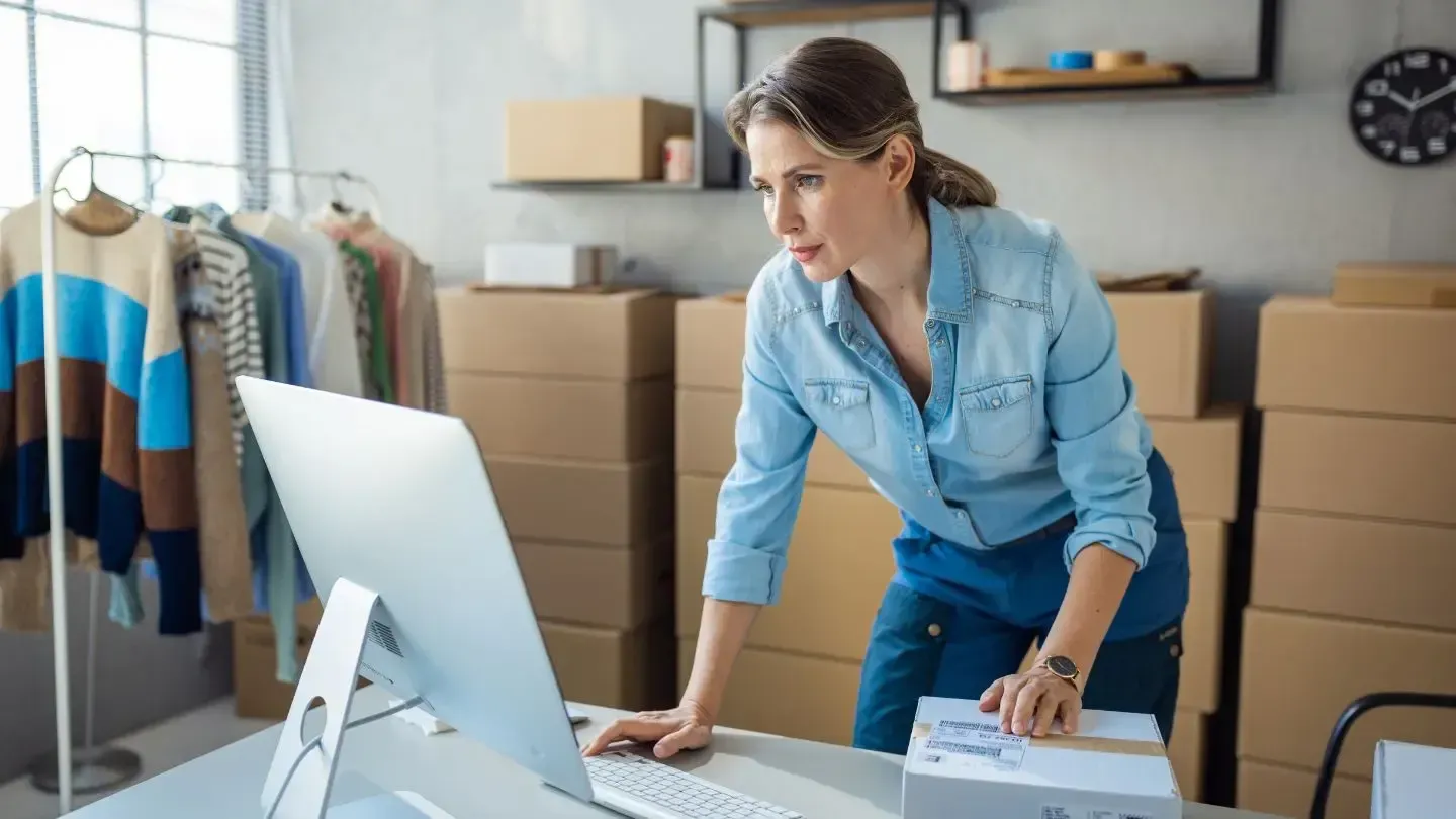 Woman in a denim shirt packages an item at a computer, surrounded by stacked shipping boxes and clothing.
