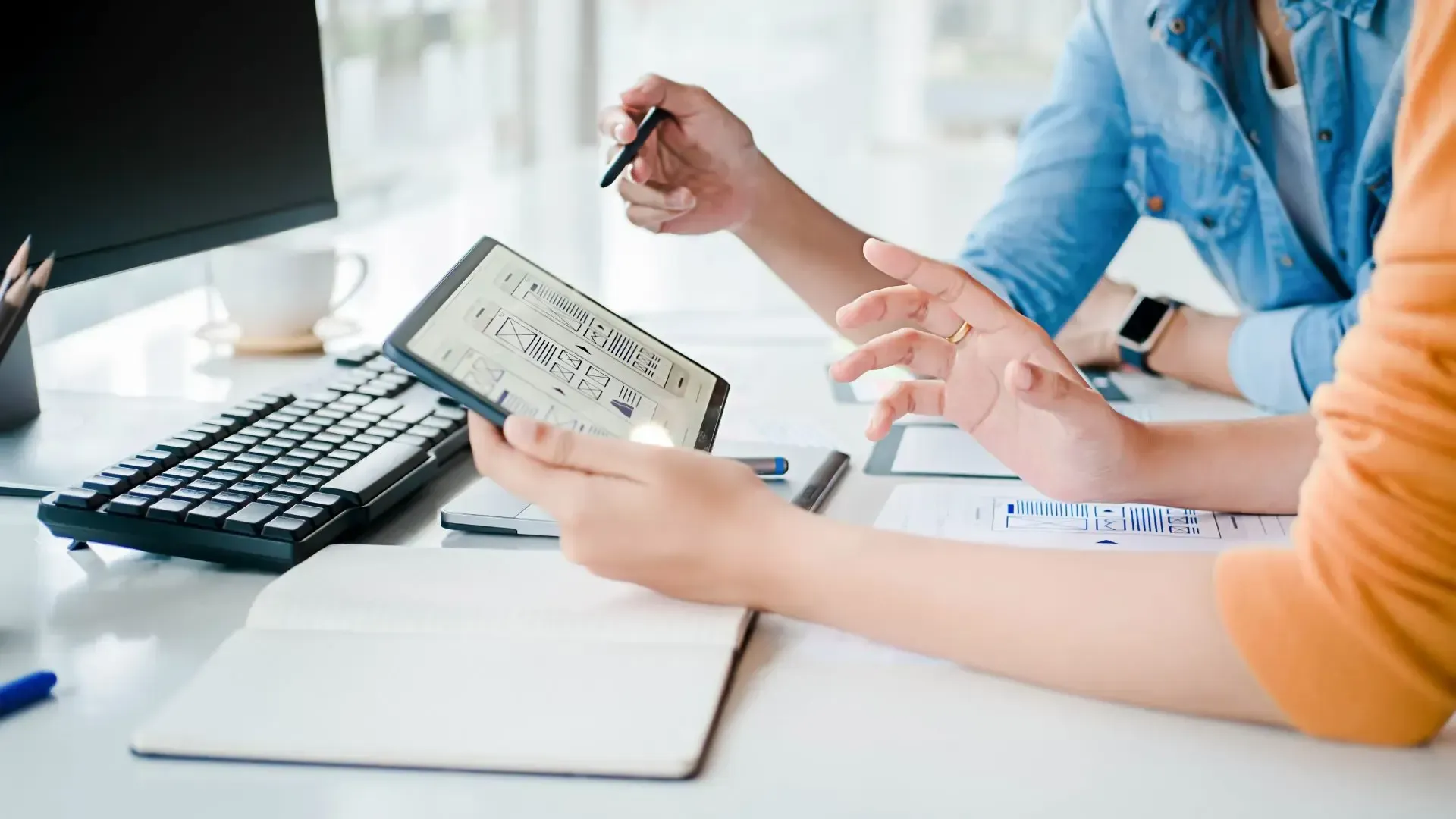 Two people at a desk reviewing data on a tablet, one pointing, and papers and keyboard nearby.