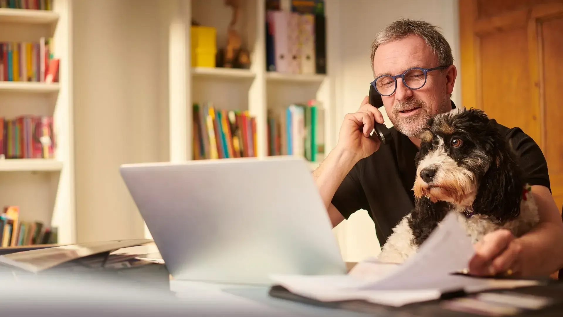 Man on phone works from home with dog, laptop, papers, bookshelves in background.