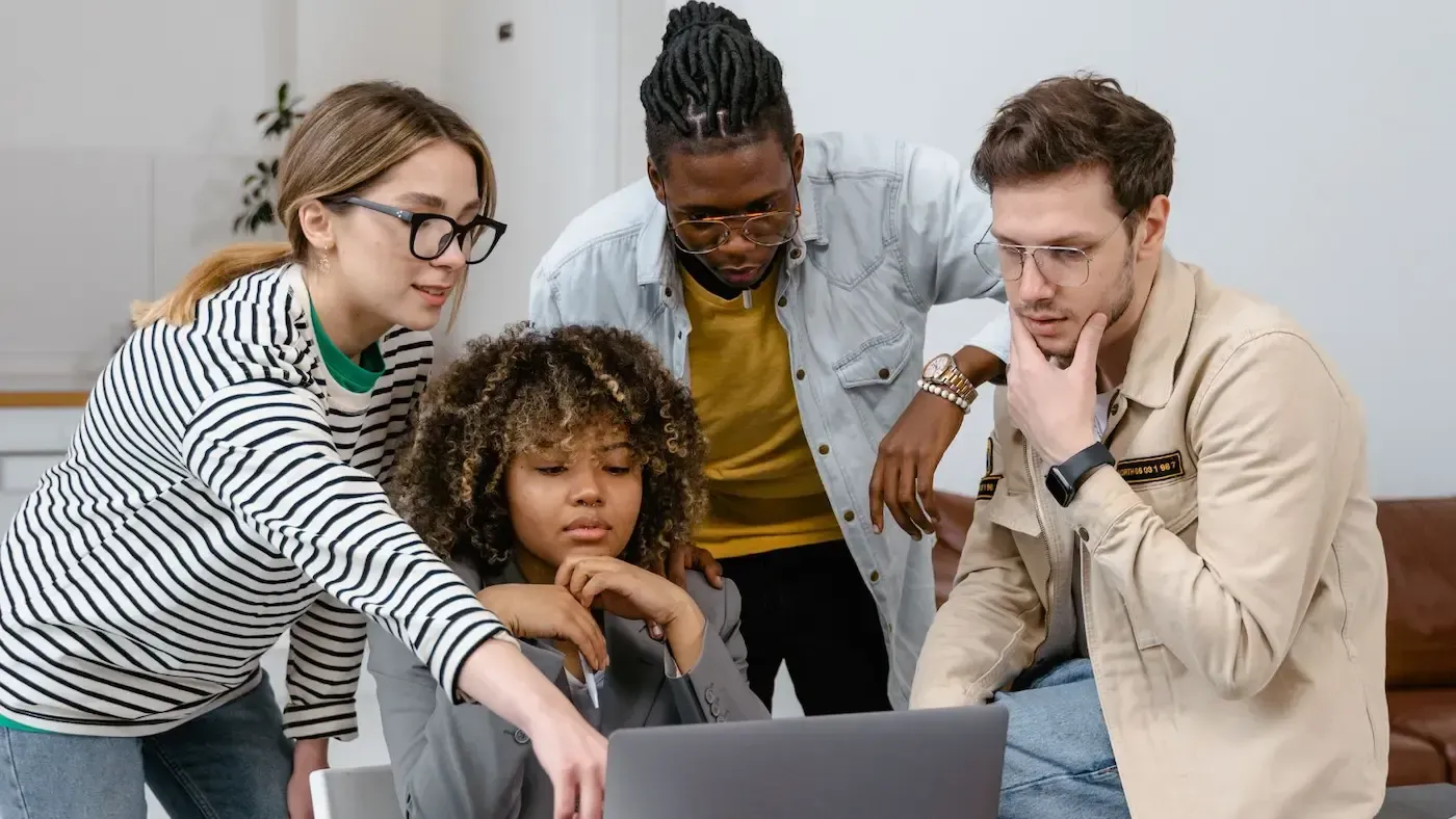 Four diverse people looking at laptop, discussing. Woman points. Indoors.