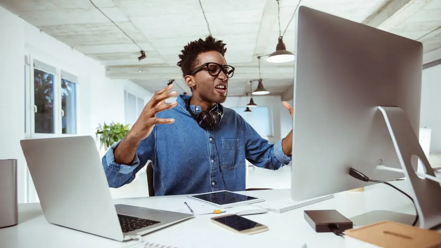 Man gesturing excitedly in front of computer, laptop, and phone at desk.