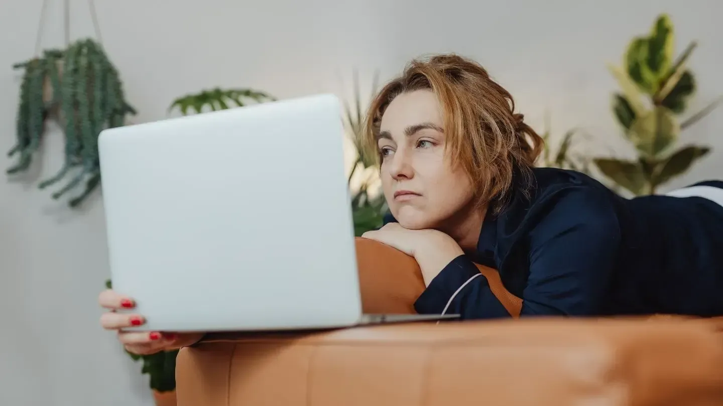 Woman using laptop, lying on a brown couch, with a serious expression. Plants in background.