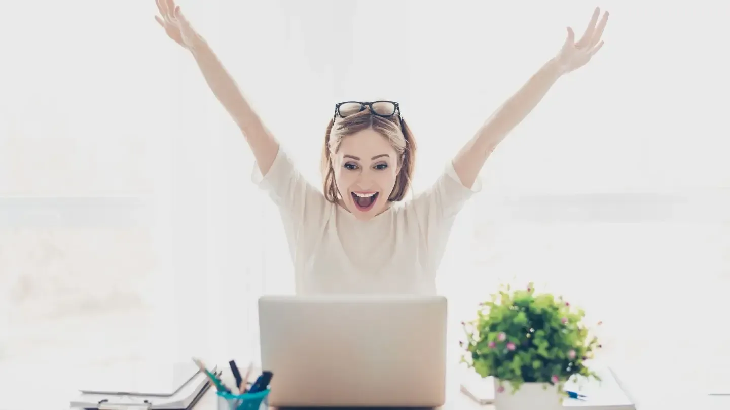 Woman at desk, arms raised in excitement, looking at laptop. White top, glasses, plant, pens.