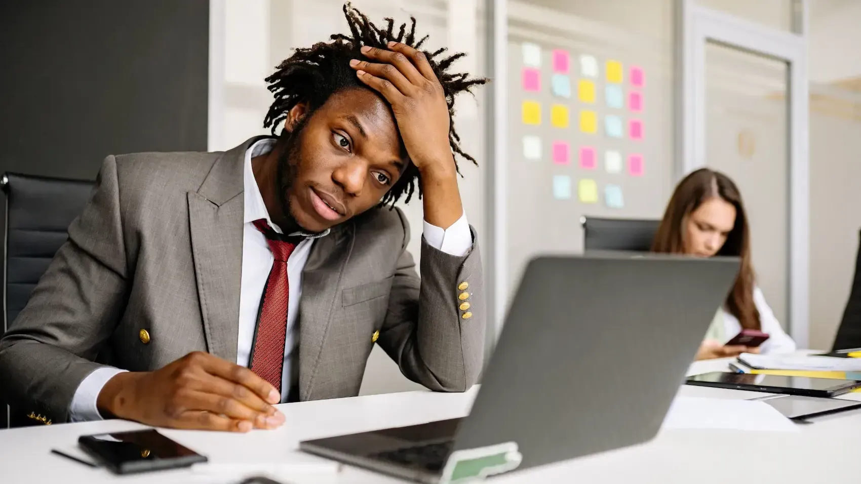 A stressed professional sits at a desk looking at a laptop with a hand on their head, while a colleague works in the back.