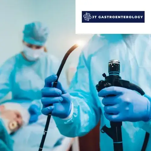 Medical staff in blue scrubs and gloves hold an endoscope in a clinical setting, with a patient visible in the background.