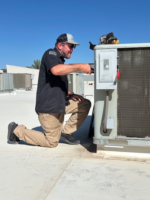 HVAC technician kneels on rooftop, inspecting a unit, holding a tool. Bright sunny day, blue sky.