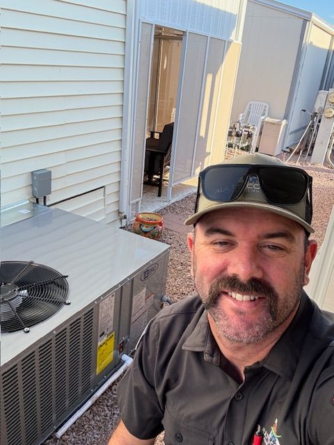 Man smiles next to an air conditioning unit; outside. Light-colored siding and a building are visible in the background.