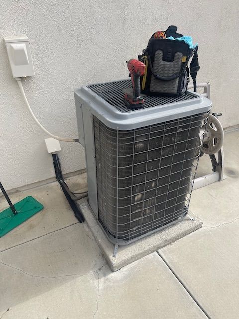 AC unit on concrete base, with tools and a tool bag on top. A brush and electrical outlet are visible.