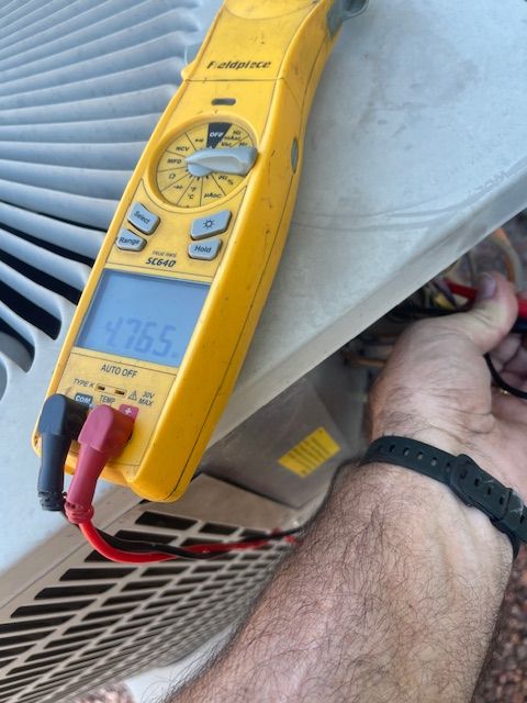 A person testing an air conditioning unit with a yellow multimeter. The display reads 476.5.