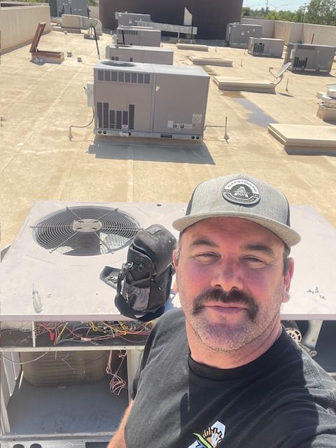 Man on a rooftop, working on an AC unit, smiling. Gray hat, black bag. Other units in the background.