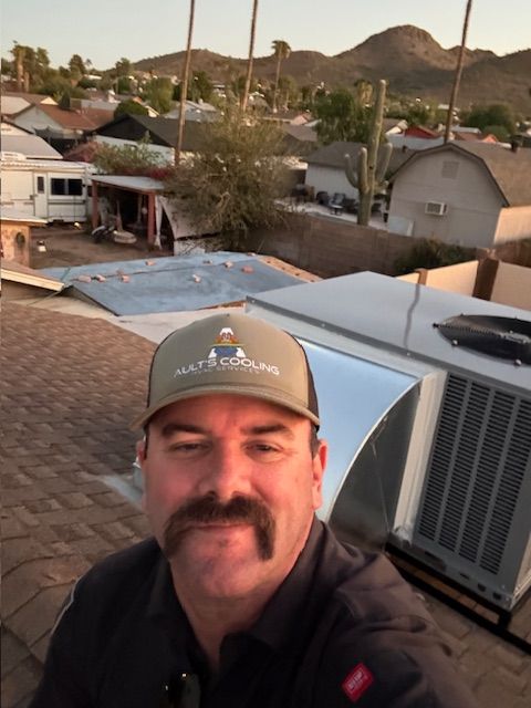 Man on a rooftop wearing a hat, smiling at the camera, with a background of houses and mountains.