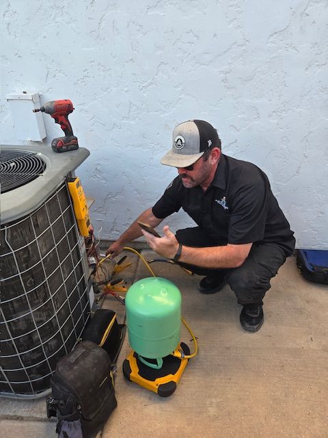 HVAC technician in cap squats near AC unit, inspecting equipment and using a phone. Outdoors.