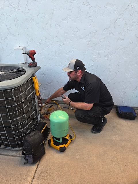 HVAC technician kneeling near an air conditioning unit, inspecting it. He wears a cap and uses tools, outdoors.