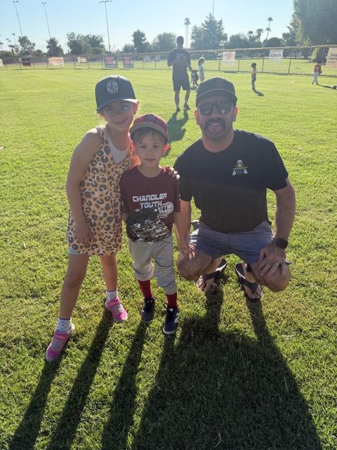 A man kneels with a girl and boy on a baseball field on a sunny day.