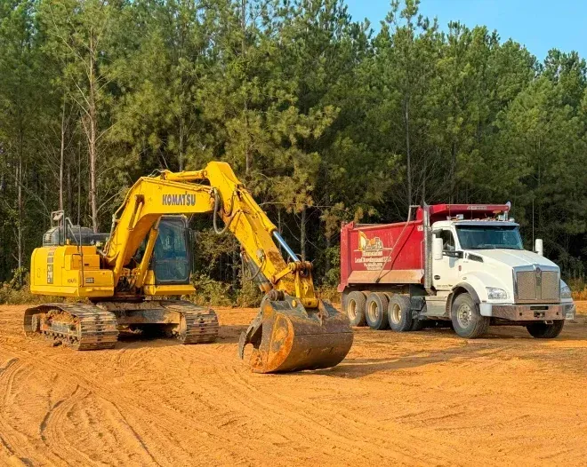 A yellow excavator and a red dump truck are parked in a dirt field.